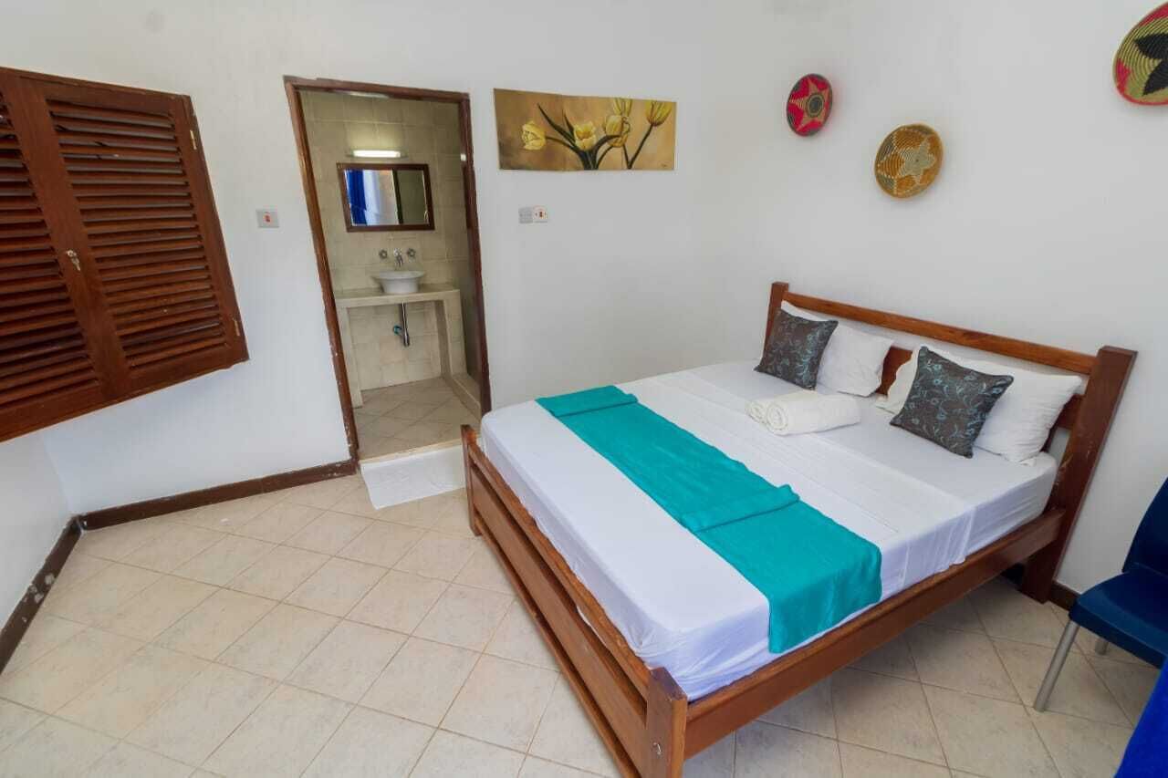 A bedroom with a wooden bed, white bedding, a teal throw, and a bathroom visible through an open door.