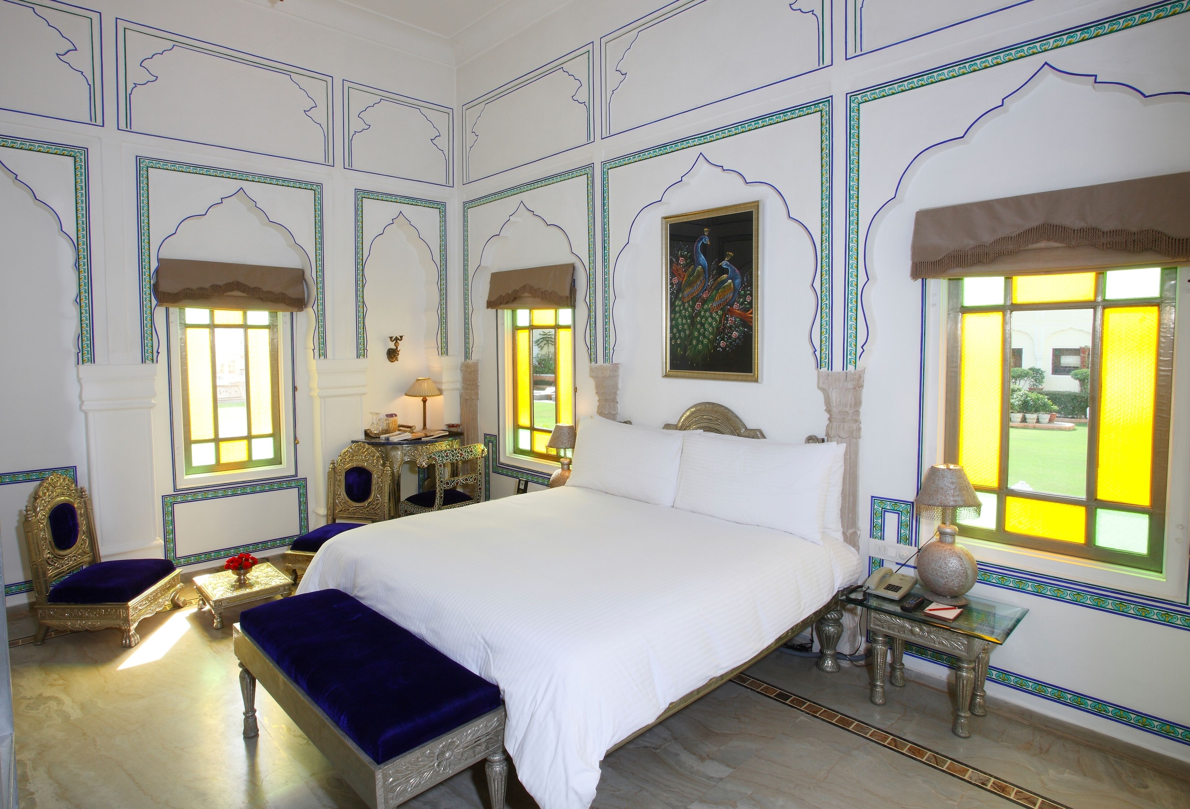 A traditional room with a large bed, two chairs, a small table with a lamp, and a decorative wall painting.