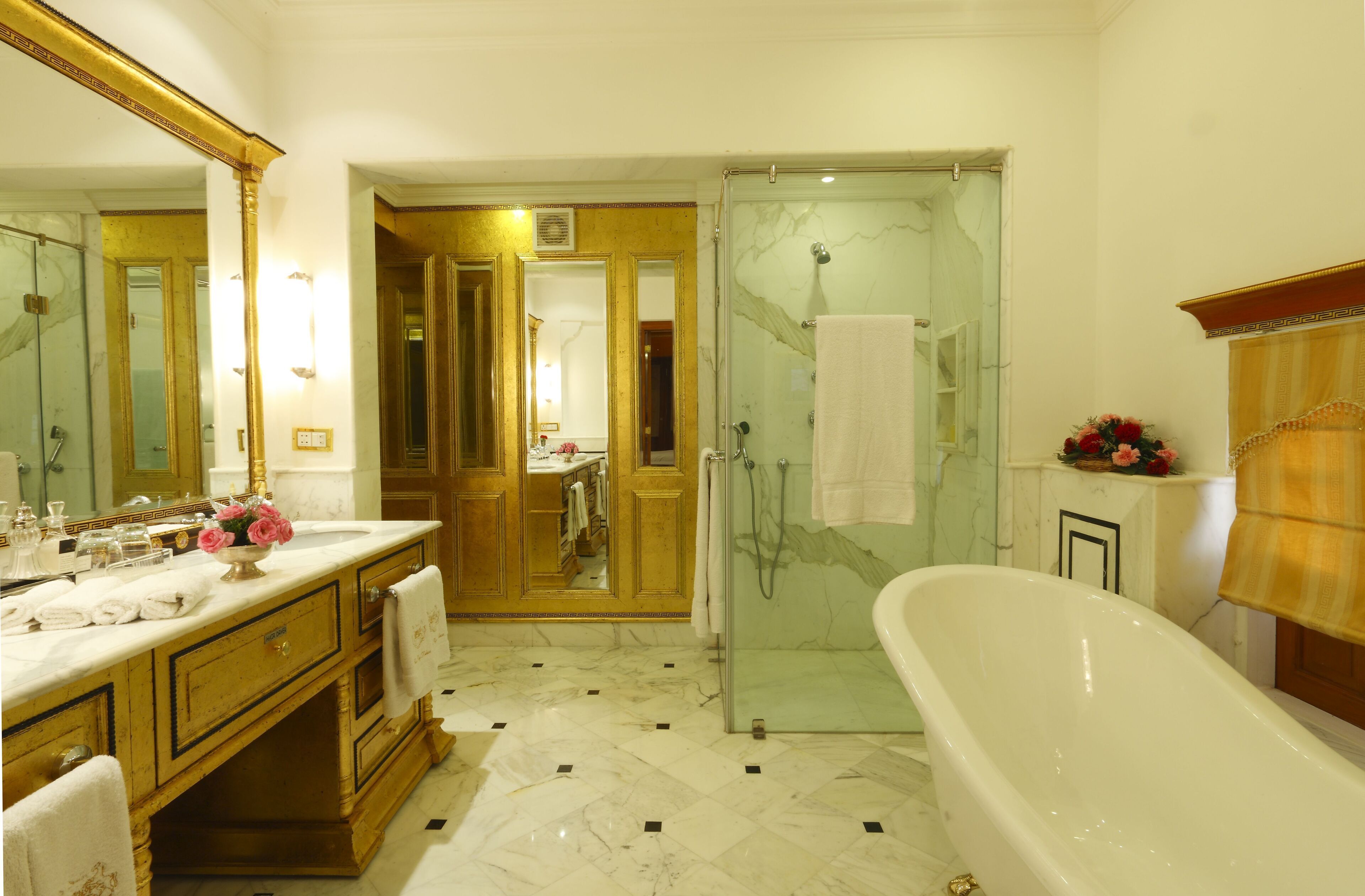 Um banheiro luxuoso com piso de mármore, uma banheira grande e independente, chuveiro envidraçado e toucador com acabamento dourado.