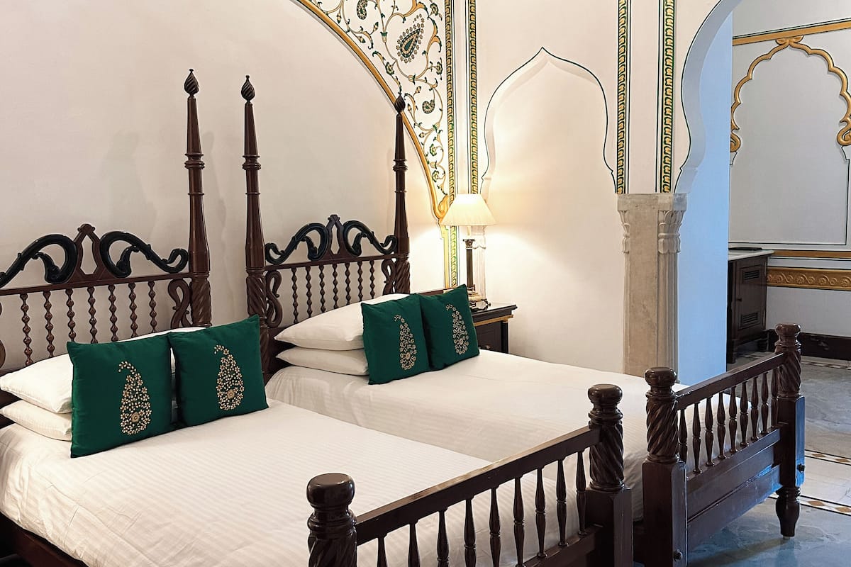 A bedroom with two beds, ornate wooden headboards, green decorative pillows, a white bedspread, and a marble floor.