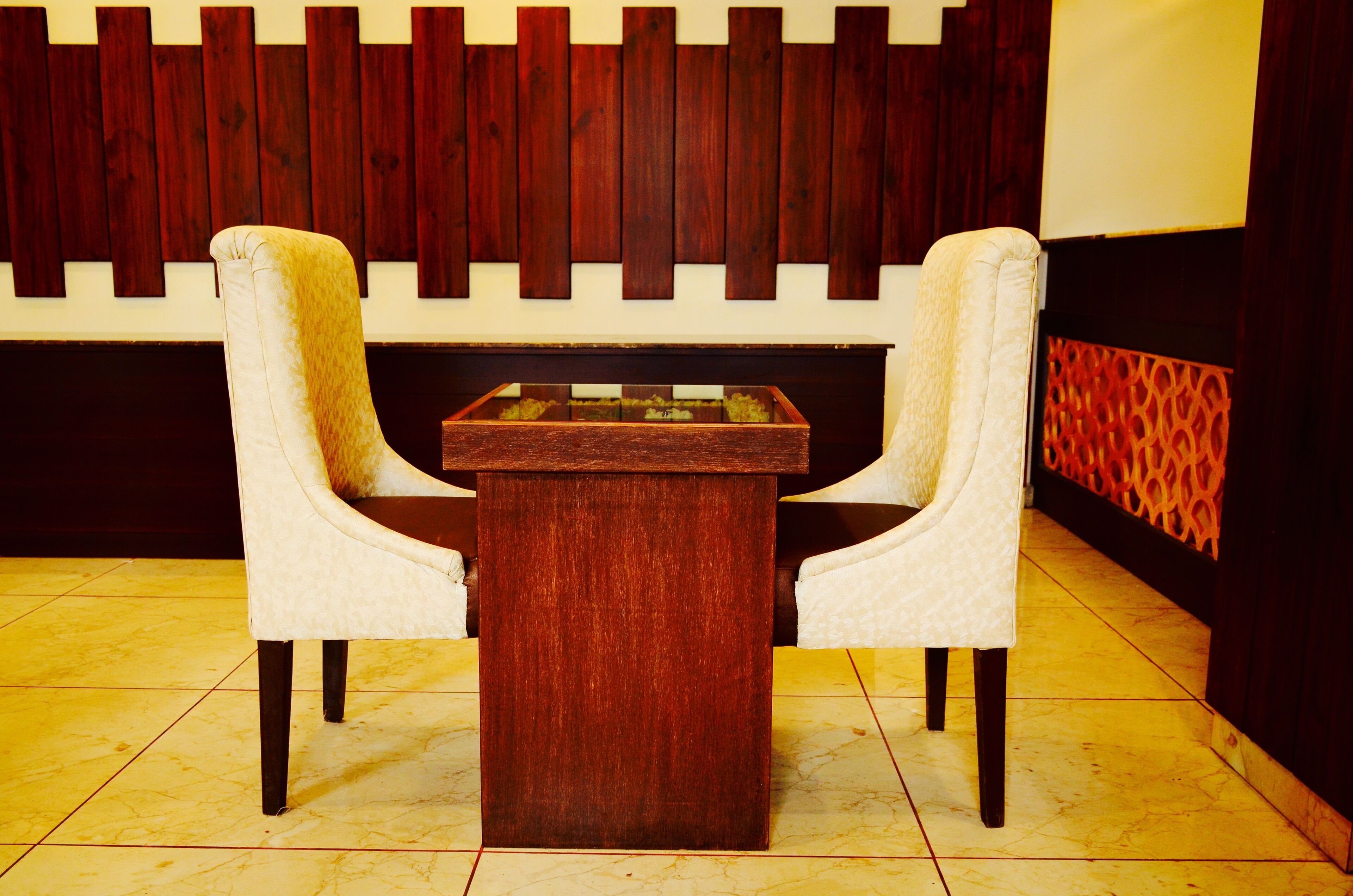 A room with a wooden wall panel, a glass-top table, and two upholstered chairs.