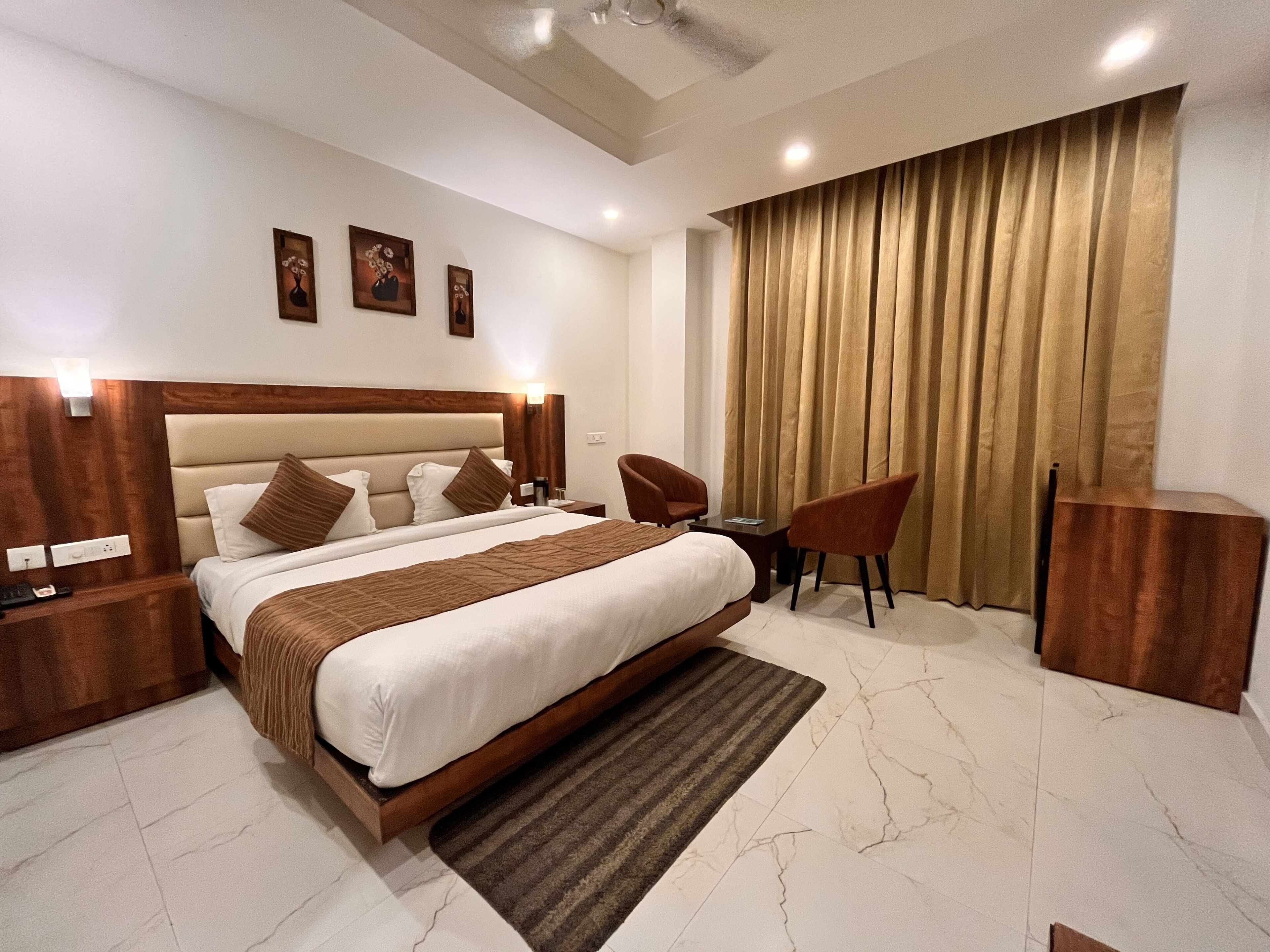 Una habitación de hotel moderna con una cama grande, un escritorio, dos sillas, un cabecero de madera y un amplio ventanal con cortinas.