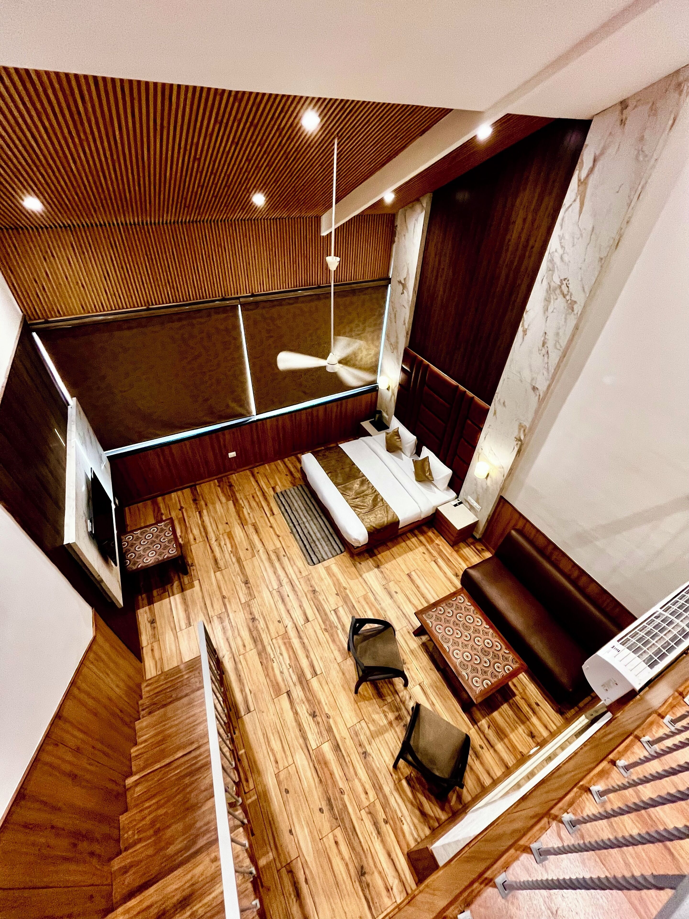 Ein modernes Hotelzimmer mit Holzboden, einem Bett, einer Sitzecke mit Sofa und Sesseln, einem Deckenventilator und einem großen Fenster.