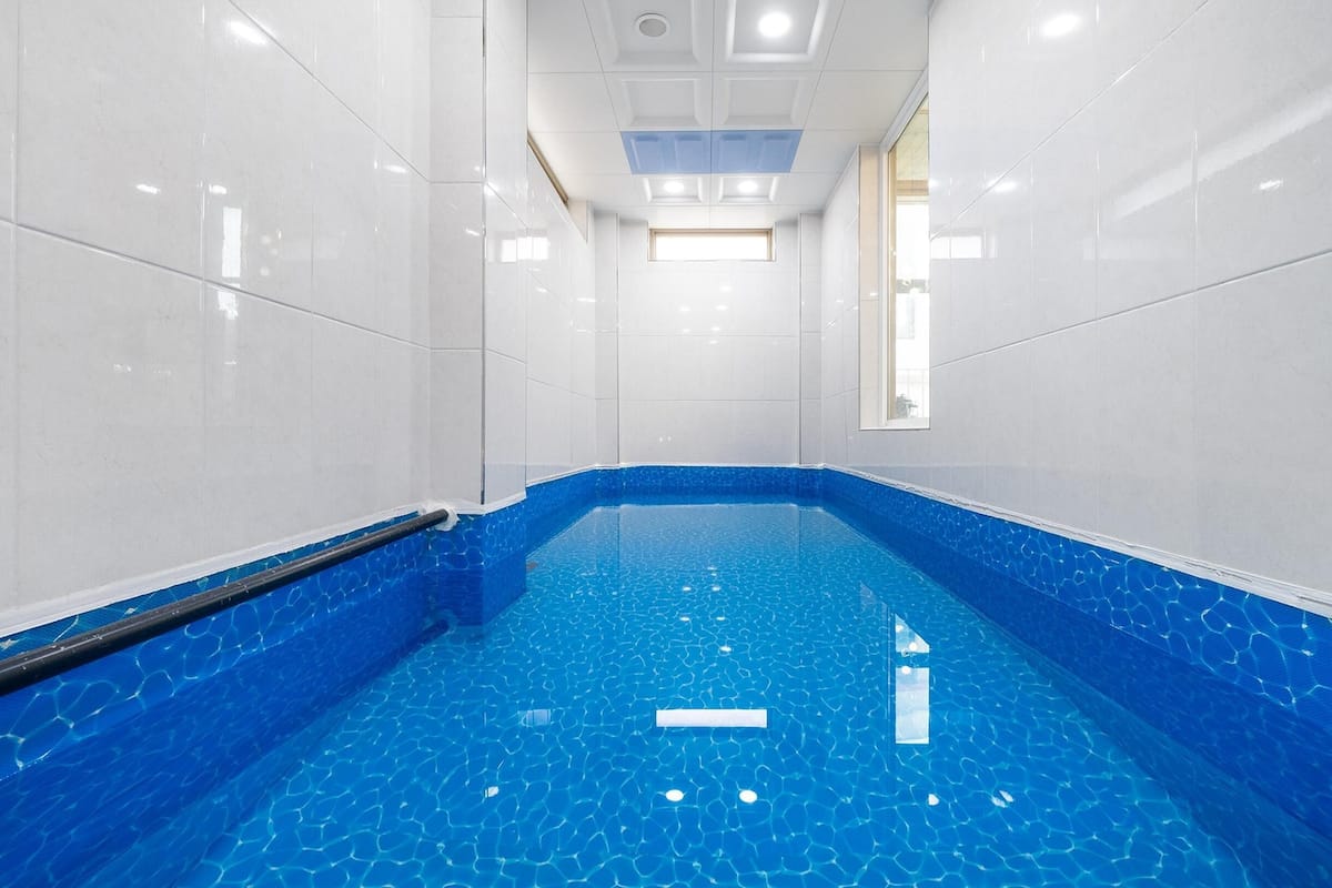 Una piscina con agua azul cristalina, rodeada de paredes de baldosas blancas y una escalera de piscina negra.