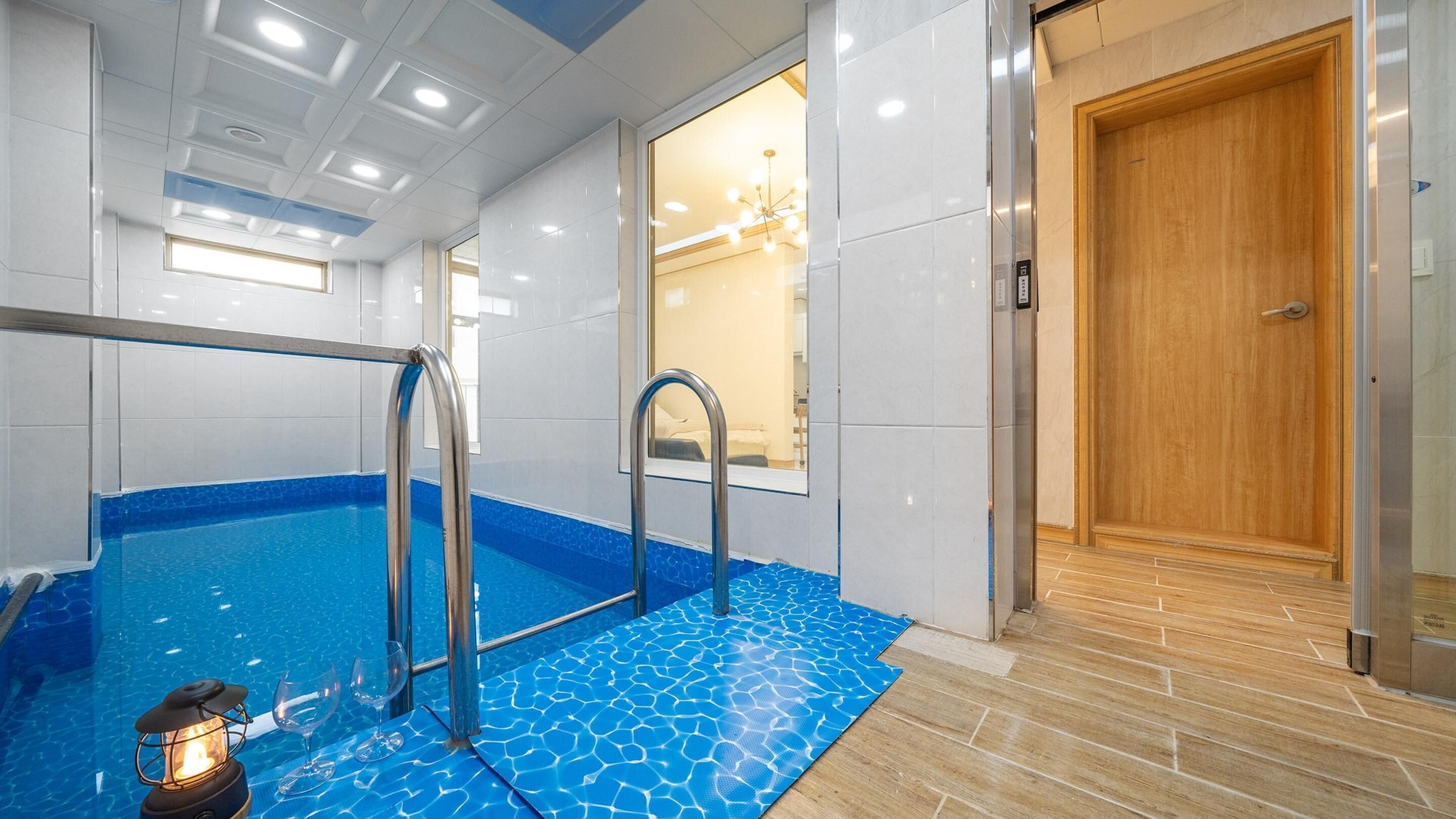 A modern indoor pool area with clear glass walls, a wooden door, and a blue tiled floor.