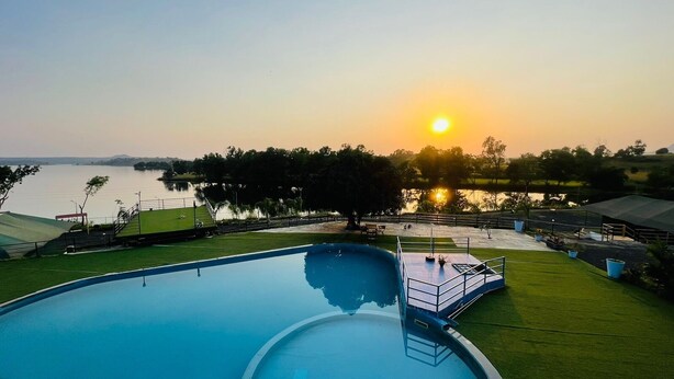 Ein Schwimmbecken mit Blick auf den Sonnenuntergang über einem Gewässer, umgeben von Grünflächen und einigen Gebäuden.