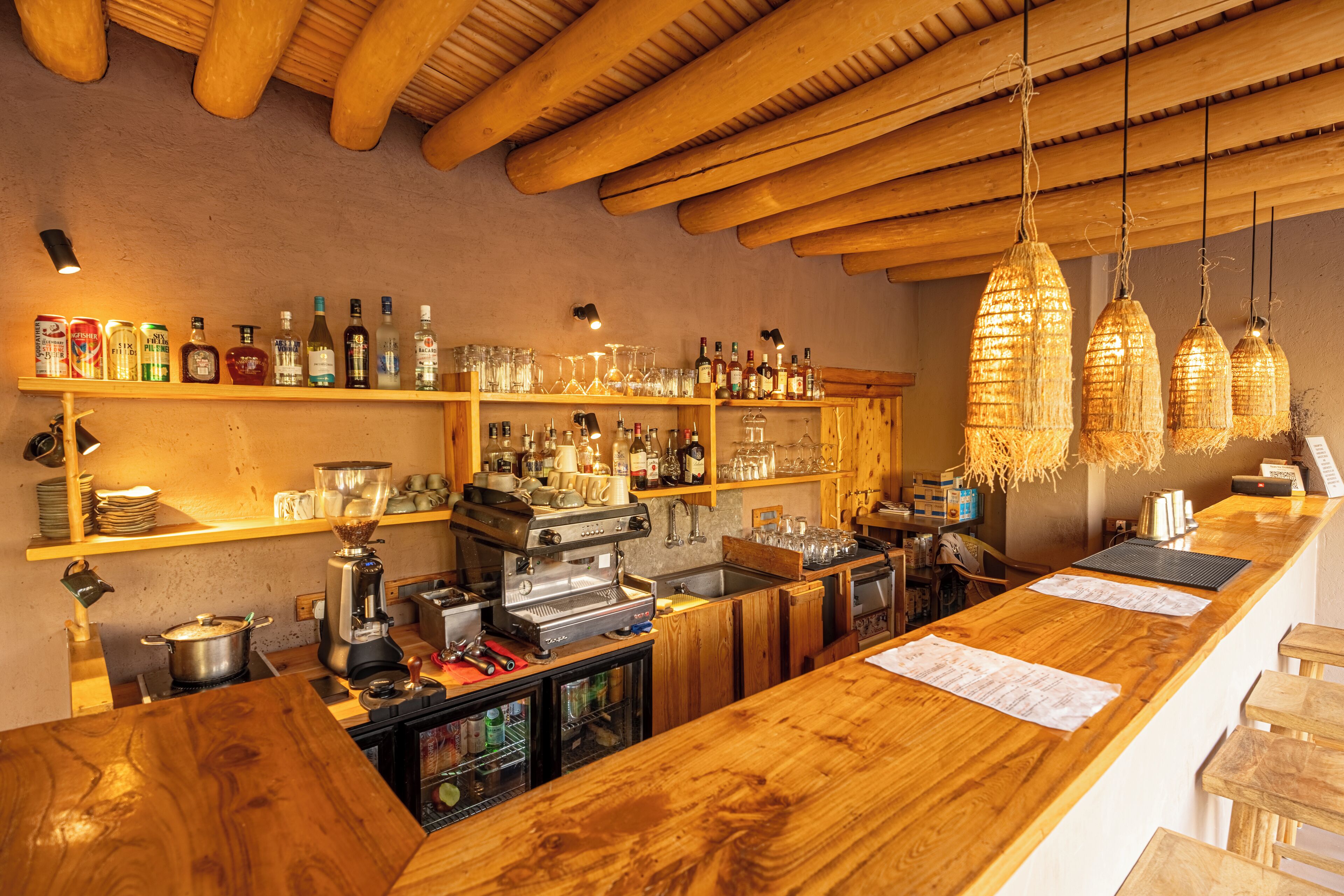 Zona de bar con estanterías de madera llenas de botellas, una máquina de café y lámparas colgantes.