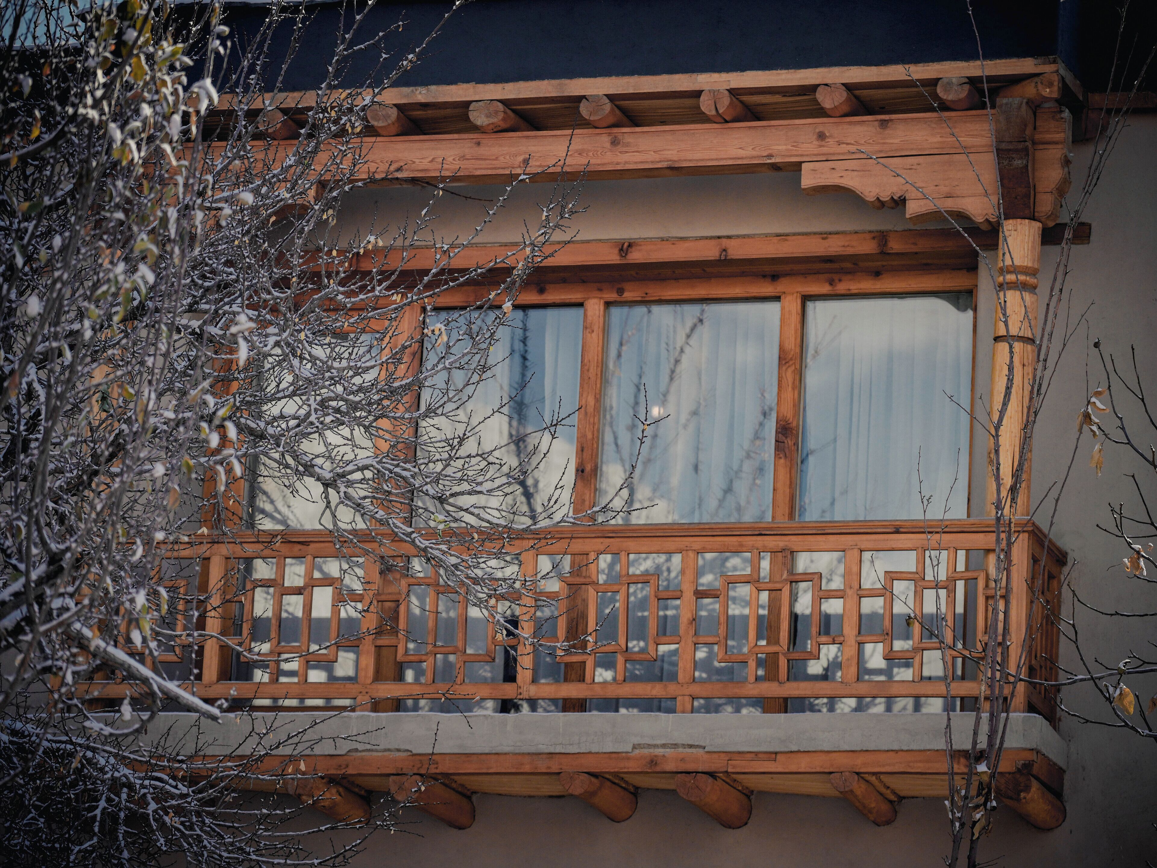 Un balcon en bois orné d’une balustrade au motif géométrique, une fenêtre donnant sur le ciel et une structure en bois au-dessus de la fenêtre.