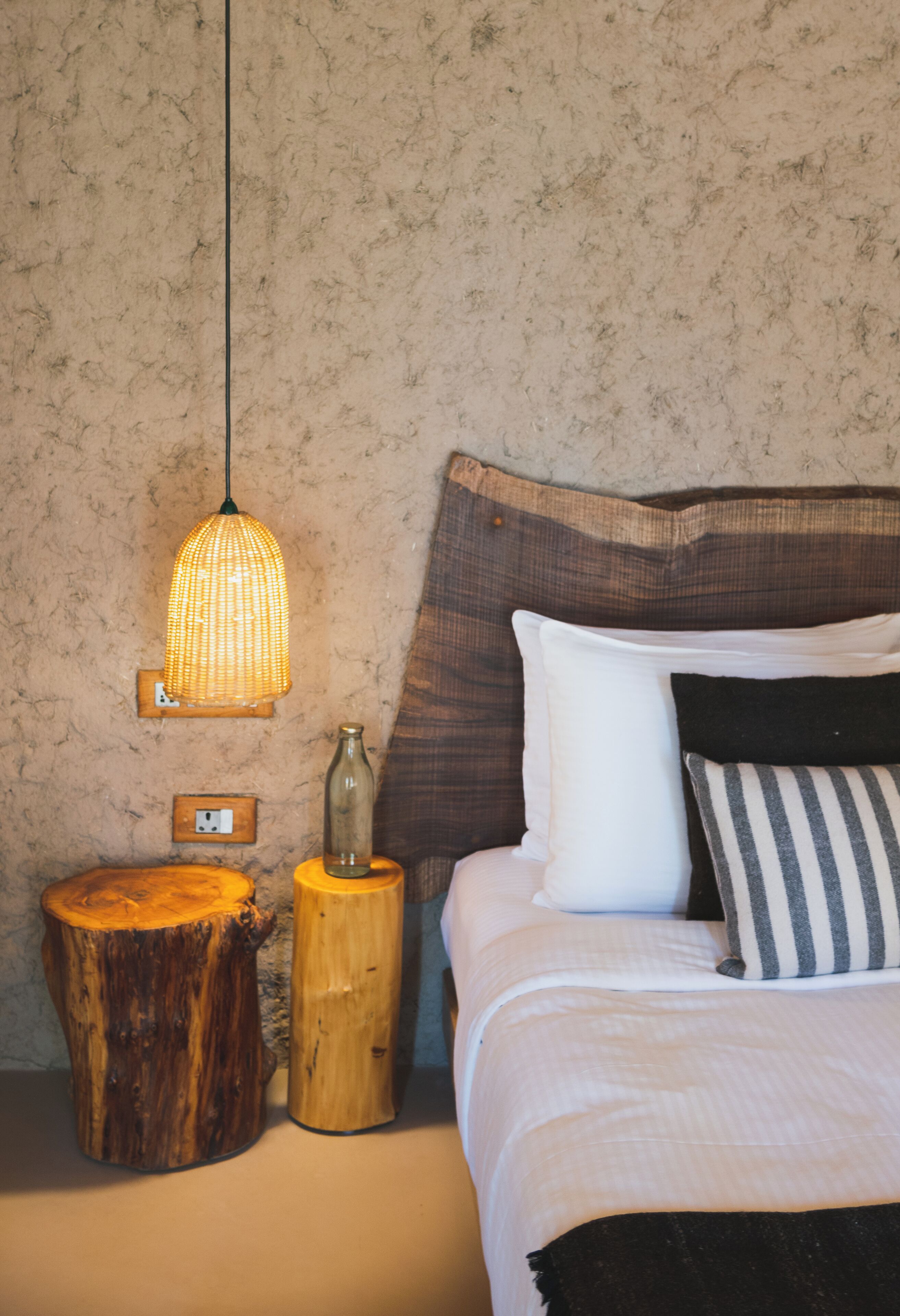 Un lit bien fait, avec des oreillers blancs et rayés, une table de chevet en bois sur laquelle se trouvent une bouteille et une lampe, et un mur à la texture intéressante.