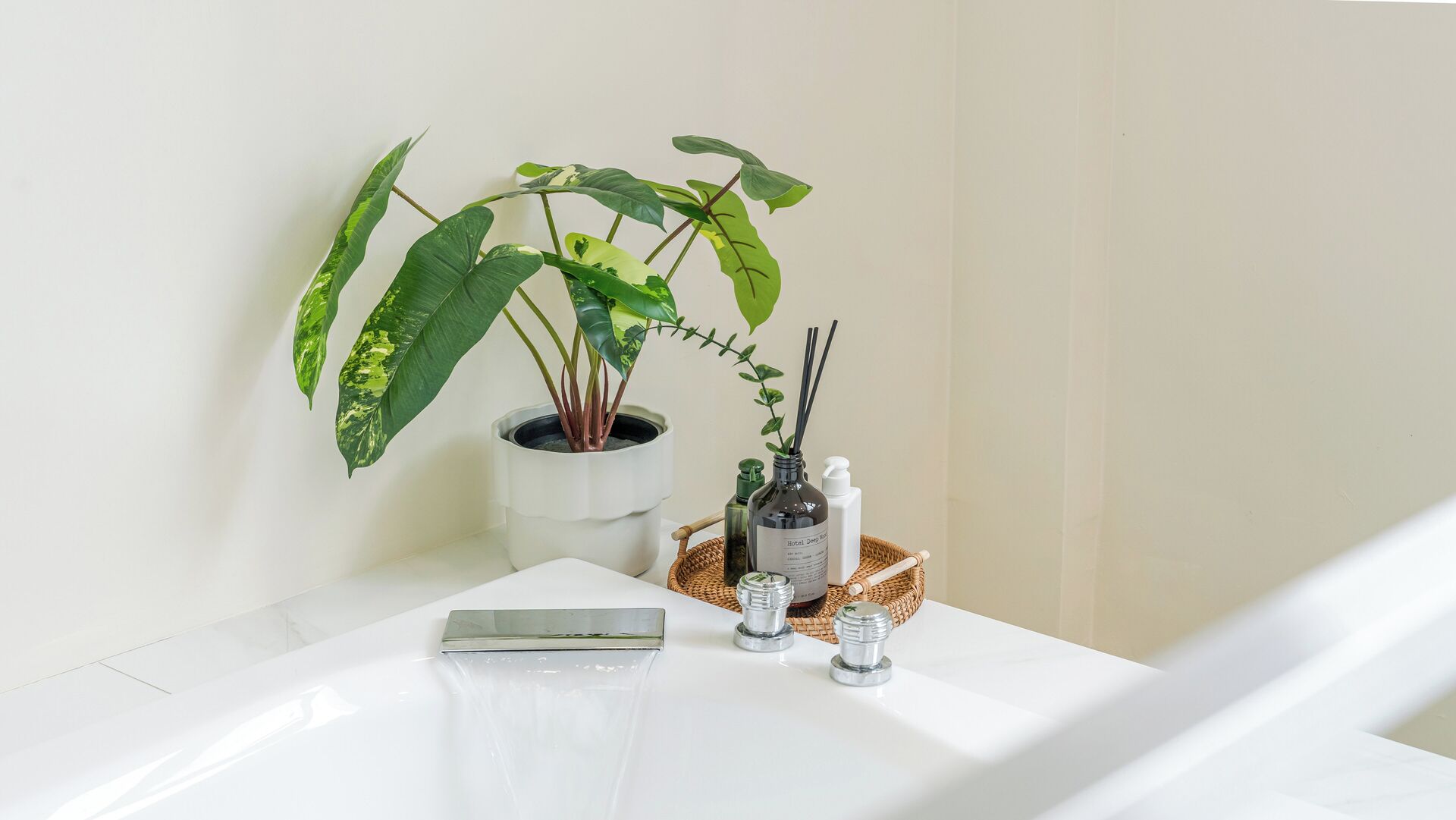 A white bathroom with a potted plant, a bottle of Hotel Deep Blue, and a small bottle on a woven basket.