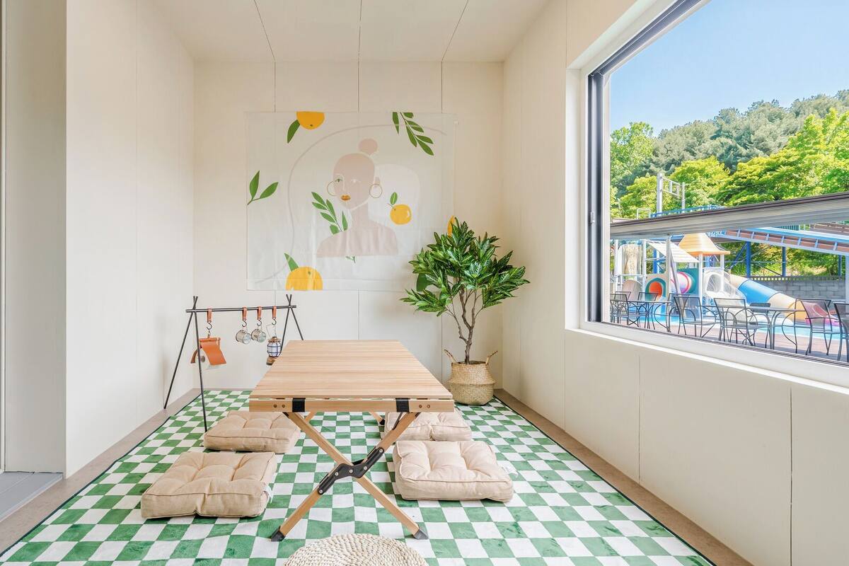 Ein Zimmer mit Schachbrettboden, einem Holztisch, Kissen, einer Hängeschaukel, einem Blumentopf und Blick auf einen Wasserpark durch ein Fenster.
