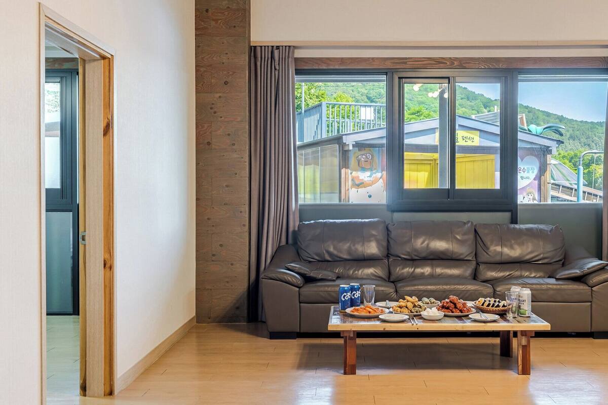 Ein Zimmer mit einer Ledersofa, einem Holztisch mit Speisen und einem großen Fenster mit Blick auf ein Gebäude und Grünflächen.