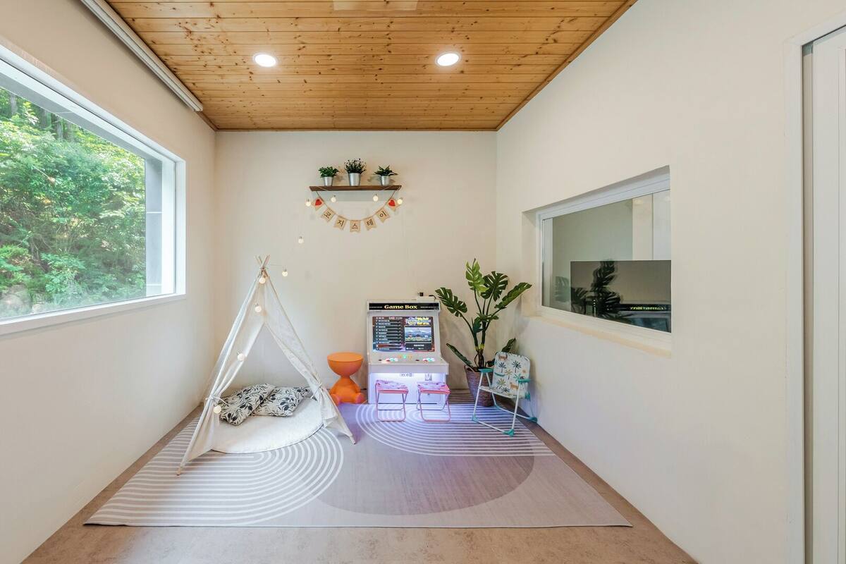 Ein Zimmer mit Holzboden, einem großen Fenster, einer GameBox, einem weißen Stuhl, einem kleinen Tisch und einer Pflanze.