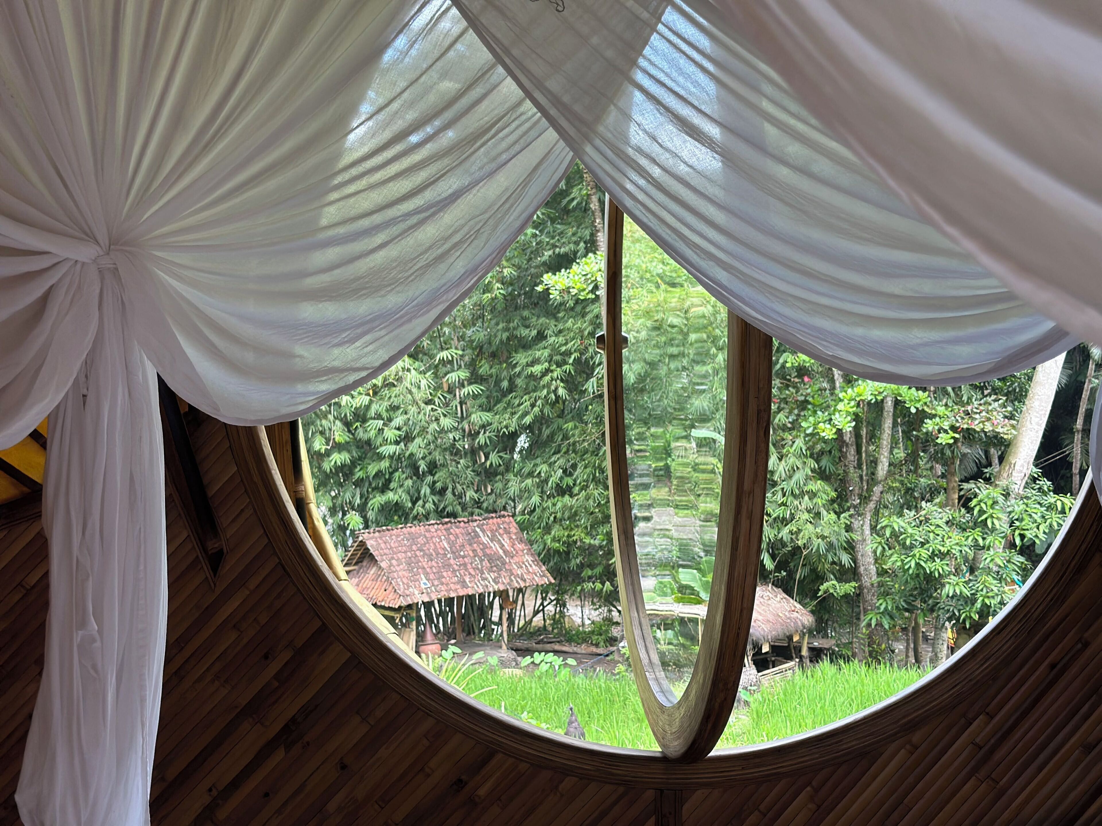 A round window with white drapes, wooden beams, and a view of a rural area with greenery and a small house.