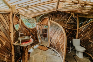 A rustic, bamboo-walled room with a stone floor, a stone basin with a soap dispenser, a woven chair, and a toilet.