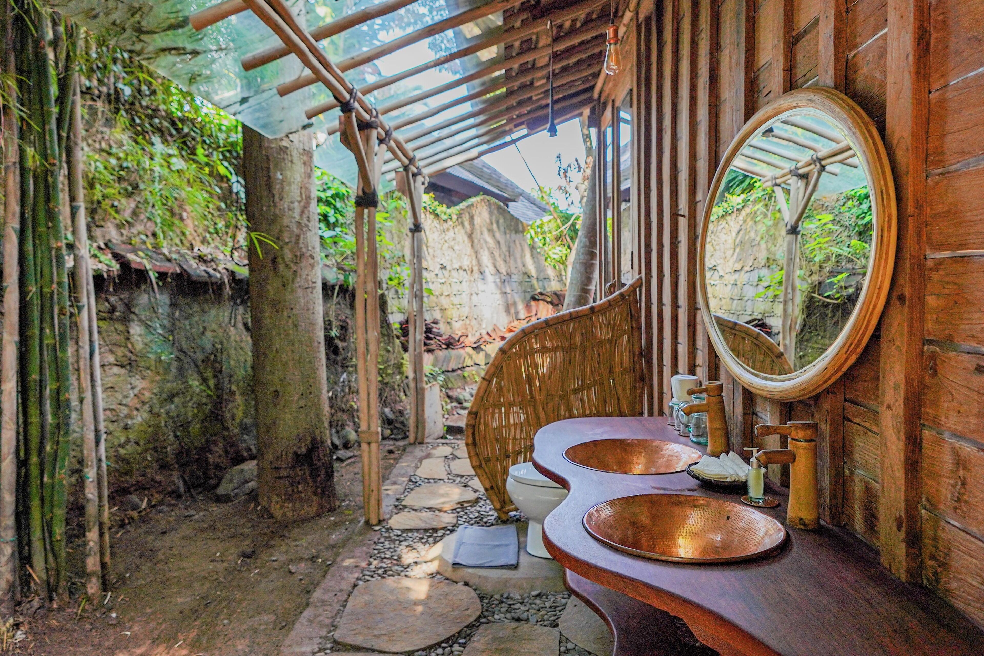 Une salle de bain rustique avec un banc en bois, un grand miroir et un chemin de pierre menant à l’extérieur.