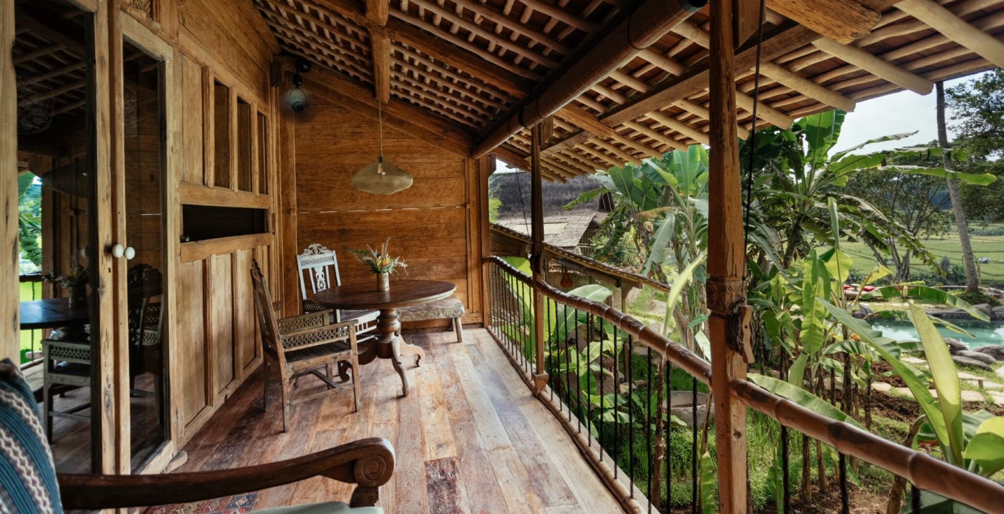 A wooden veranda with a wooden table and chairs, a hanging light fixture, and a view of a lush garden and river.