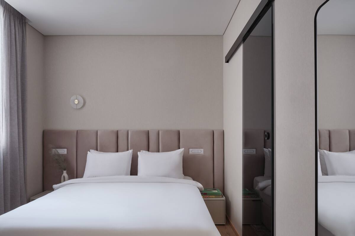 A modern hotel room with a large bed, a bedside table, a nightstand, a mirror, and a view of the bathroom.