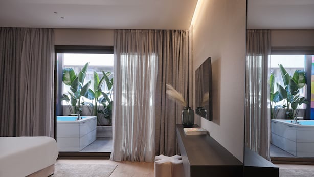 A modern hotel room with a large bed, a bedside table with a vase and book, a mirror, and a view of a balcony with plants. - Nur Outline Aparthotel (Athens)
