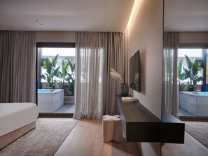 A modern hotel room with a large bed, a bedside table with a vase and book, a mirror, and a view of a balcony with plants. - Nur Outline Aparthotel (Athens)