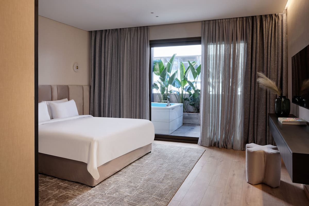 A modern bedroom with a large bed, a small bathroom, and a view of a balcony with plants.