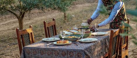 A dining table set outdoors with chairs, a person serving food, and an olive tree in the background.