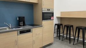 Cocina compacta equipada con lavavajillas, cafetera, microondas y horno pequeño. Hay tres taburetes negros pegados a la pared.