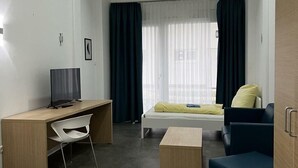 A modern hotel room with a wooden desk, a bed with a yellow pillow, a blue armchair, and a window with sheer curtains. - Fabella Flats Forchheim - 20 (Forchheim)