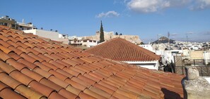 A rooftop with terracotta tiles, a white building with a red roof, and a distant view of a cityscape with historical architecture.