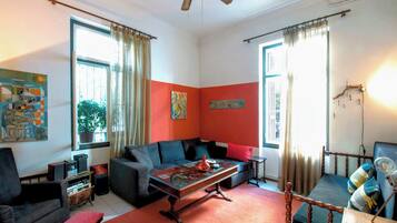 A living room with a sofa, a coffee table, and a chair. There are two windows with curtains, a ceiling fan, and artwork on the walls.