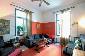 A living room with a sofa, a coffee table, and a chair. There are two windows with curtains, a ceiling fan, and artwork on the walls.