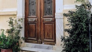 Une porte en bois à deux battants, ornée de motifs métalliques complexes, est encadrée de plantes en pot et intégrée à un bâtiment dont la façade est de couleur crème et la base en pierre.