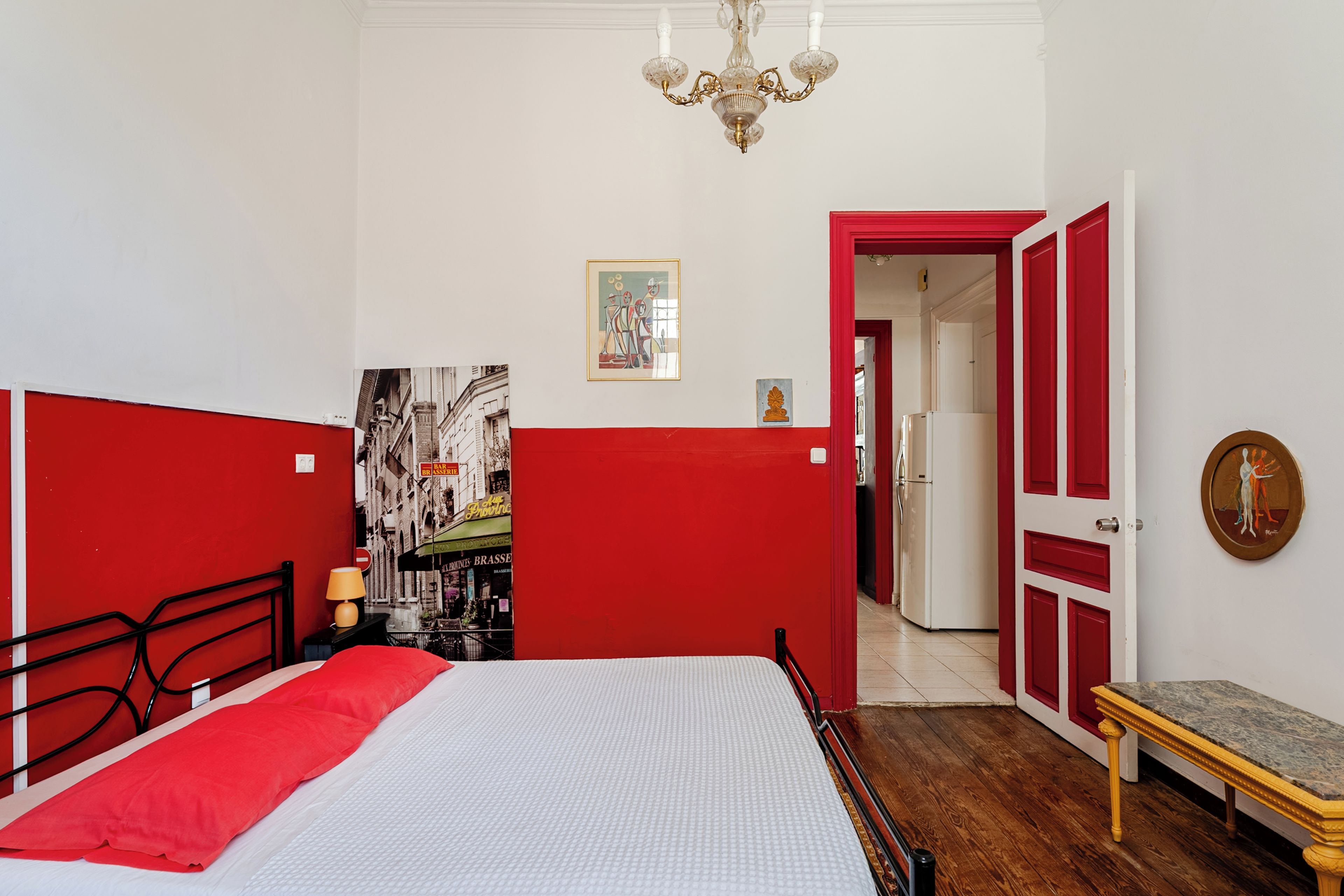 A bedroom with a bed, a nightstand, a red partition, a chandelier, a door, and a hallway leading to another room.