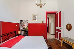A bedroom with a bed, a nightstand, a red partition, a chandelier, a door, and a hallway leading to another room.