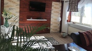 Ein modernes Wohnzimmer mit einer Wandpaneinung aus Holz, einem Flachbildfernseher, einer Pendelleuchte und einem großen Fenster mit Vorhängen.
