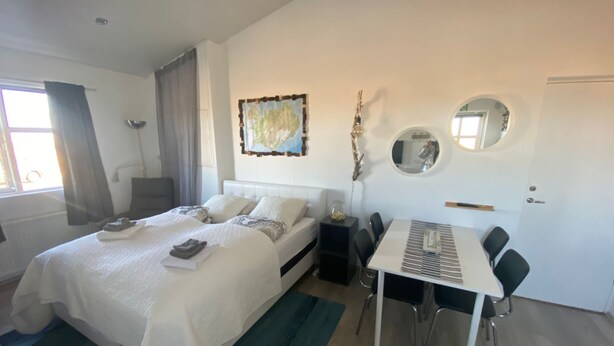 Een moderne hotelkamer met een groot bed, een eettafel met stoelen, een klein bijzettafeltje, een spiegel en een raam met gordijnen.