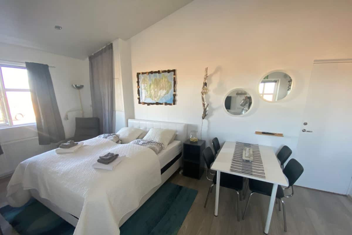 A modern hotel room with a large bed, a dining table with chairs, a small side table, a mirror, and a window with curtains.