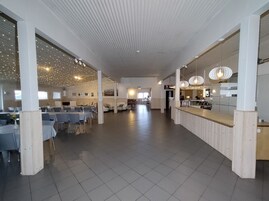 Um espaço amplo e interno, com um balcão comprido, várias mesas e cadeiras, indicando um local para refeições ou reuniões.