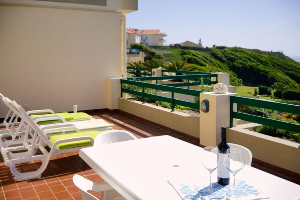 A balcony with white lounge chairs, a table, and a bottle of wine.