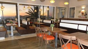 A restaurant with wooden tables and orange cushioned chairs, large windows with a view, and pendant lighting.