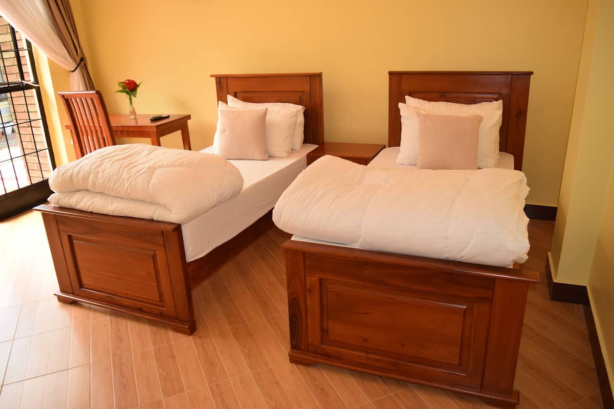 Dos camas de madera con ropa blanca, una mesita de noche de madera, una silla y un jarrón con una flor.