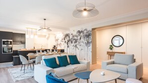 Un salon moderne avec un canapé, une table basse et des fauteuils. On y trouve également un coin repas avec une table et des chaises. La cuisine est visible au fond.