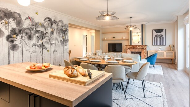 Un espace de repas moderne avec une table en bois dressée pour six personnes, un comptoir de cuisine avec du pain et des fruits, et un salon doté d’une cheminée et de canapés.