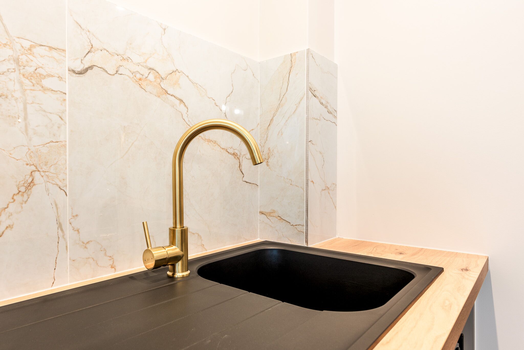 A kitchen with a black sink, gold faucet, and marble backsplash.