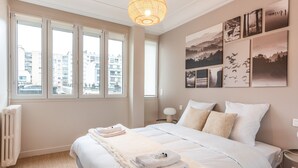 Une chambre avec un grand lit, un radiateur, une table de chevet avec une lampe et une vue sur les bâtiments par la fenêtre.