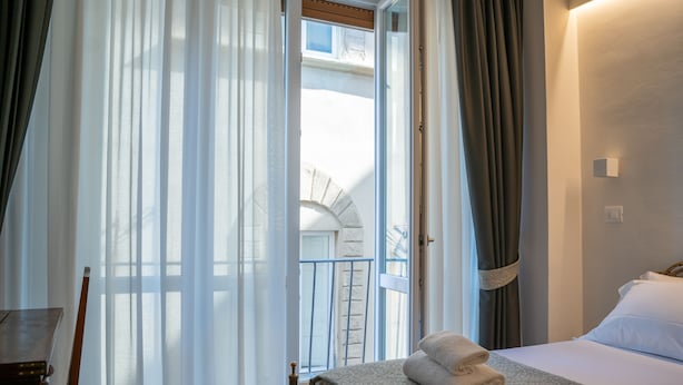 Ein Hotelzimmer mit Bett, Sessel, Tisch und Blick auf einen Balkon mit Steinmauer und Fenster.