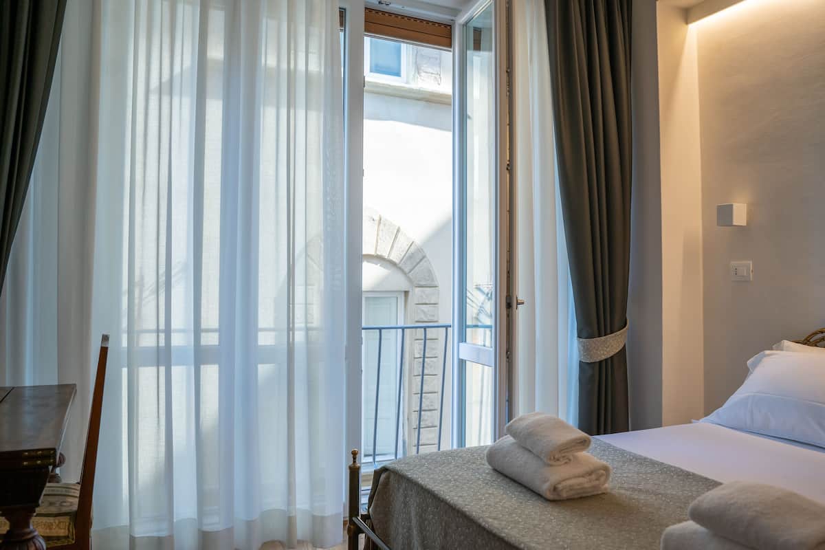 Et hotelværelse med en seng, en stol, et bord og udsigt til en balkon med en stenvæg og et vindue.