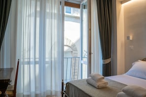 Ein Hotelzimmer mit Bett, Sessel, Tisch und Blick auf einen Balkon mit Steinmauer und Fenster.