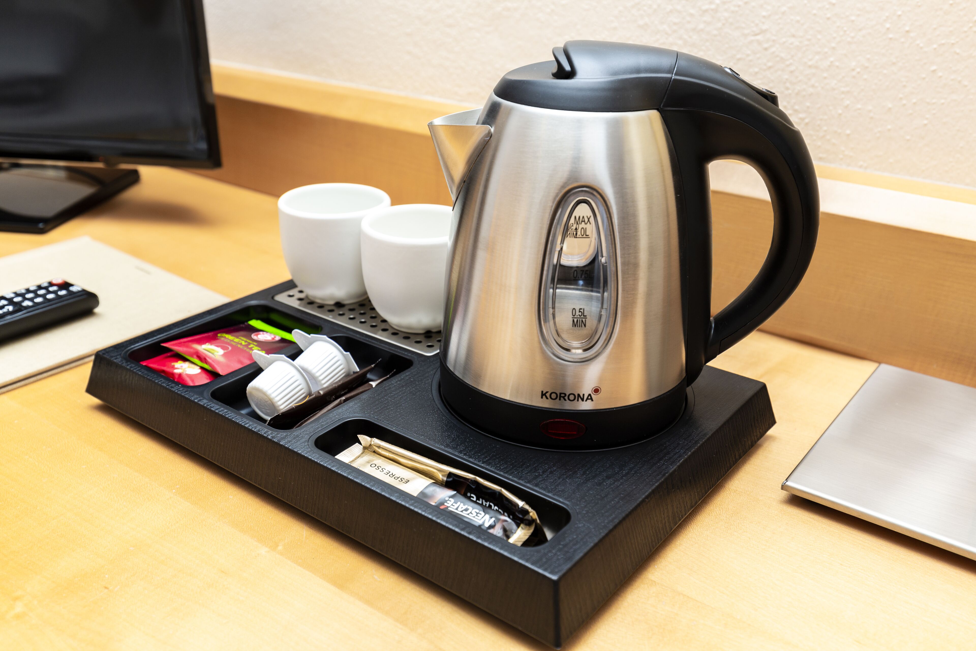 A stainless steel kettle with a black handle on a black tray, accompanied by two white cups and a remote control.