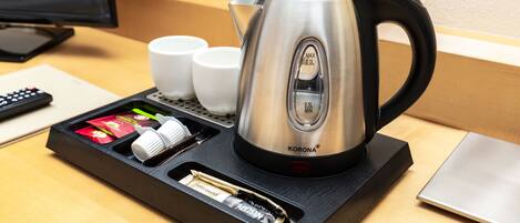 A stainless steel kettle with a black handle on a black tray, accompanied by two white cups and a remote control.